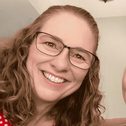 Smiling woman with curly blonde hair wearing glasses and a red patterned blouse.